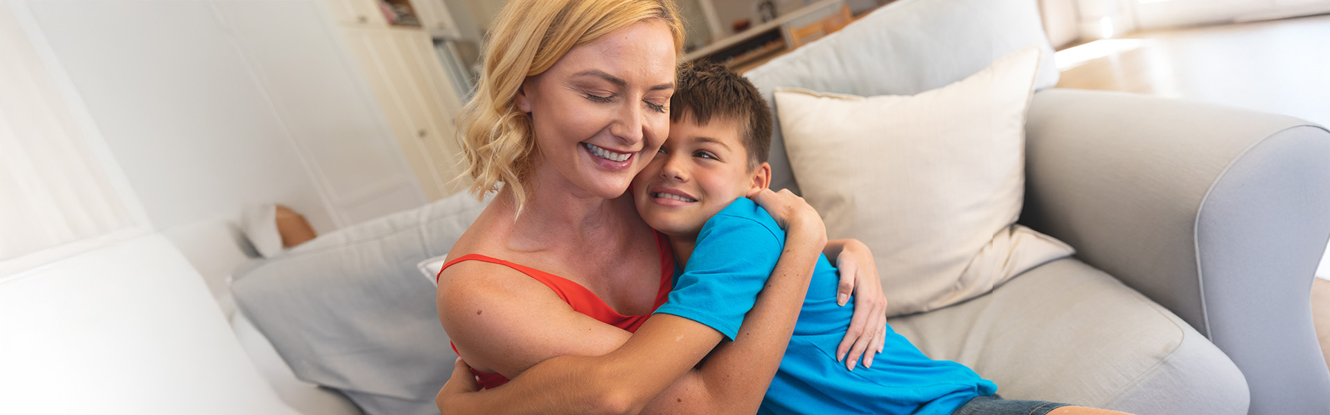 Middle aged female foster carer sits on the sofa with her foster son. They share an embrace while he is looking at her smiling