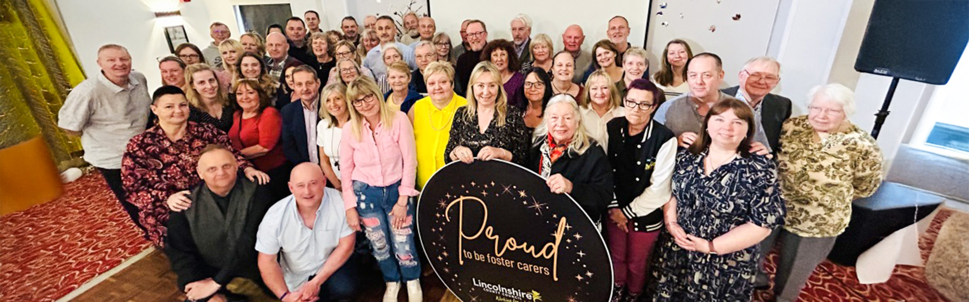 Large group of people at an awards ceremony holding a sign that says "Proud to be a foster carer" followed by the Lincolnshire County Council logo