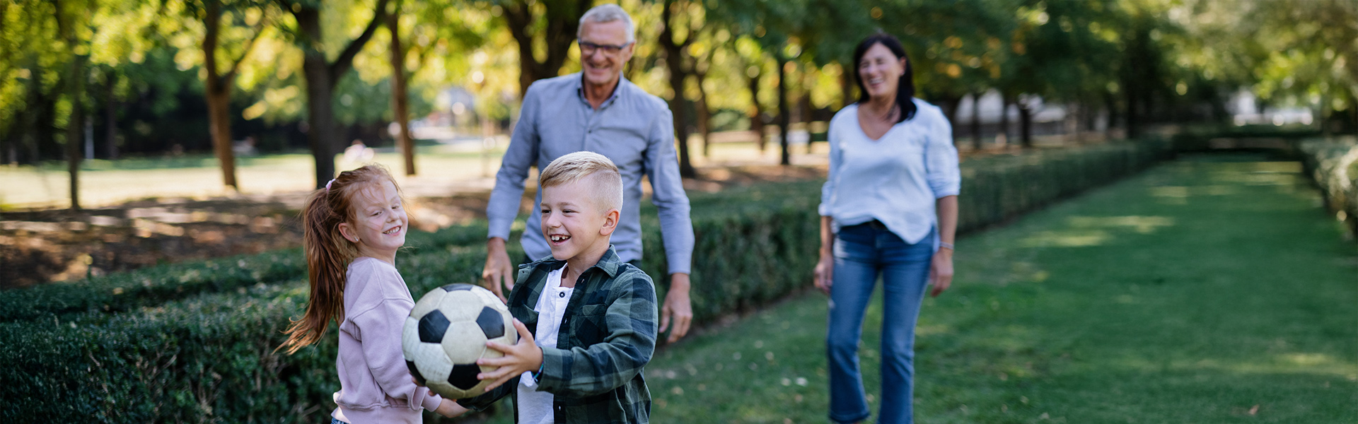 Little boy holds a football while smiling. In the background a little girl is smiling with their foster mum and dad walking towards them smiling