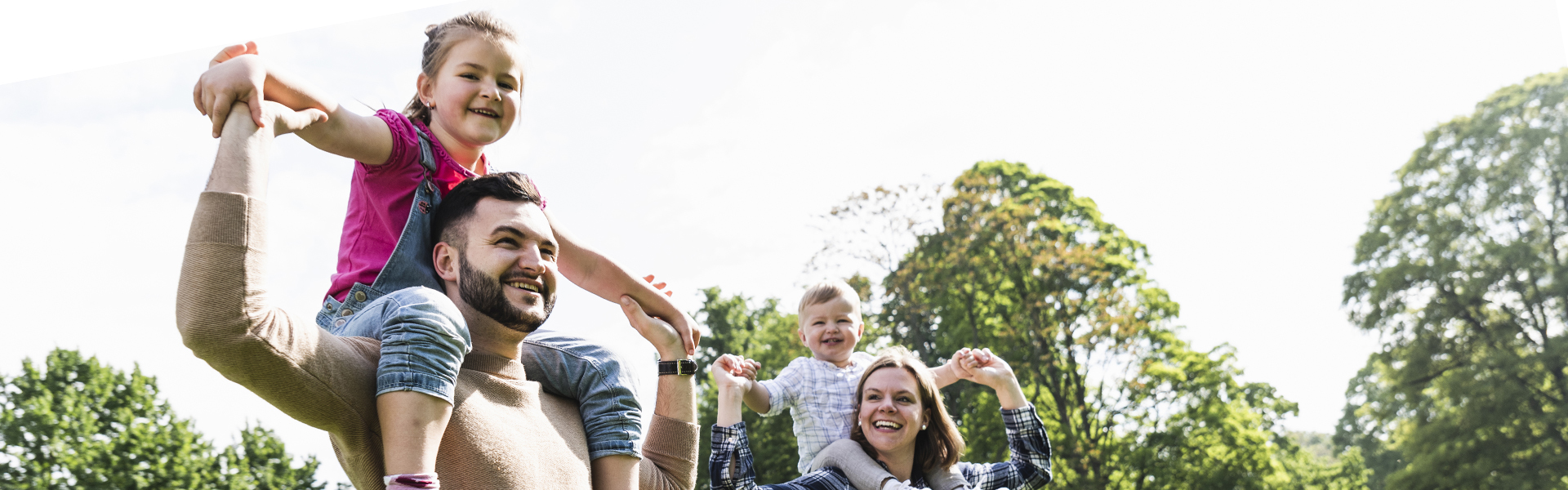Foster dad and foster mum carrying young children on their shoulders smiling while walking through the park