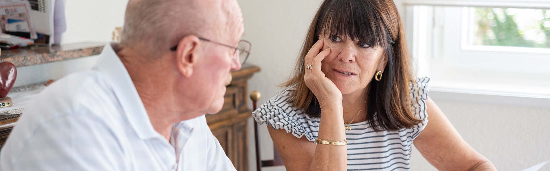 Older couple sitting at dining table holding a piece of paper and looking at each other as they discuss