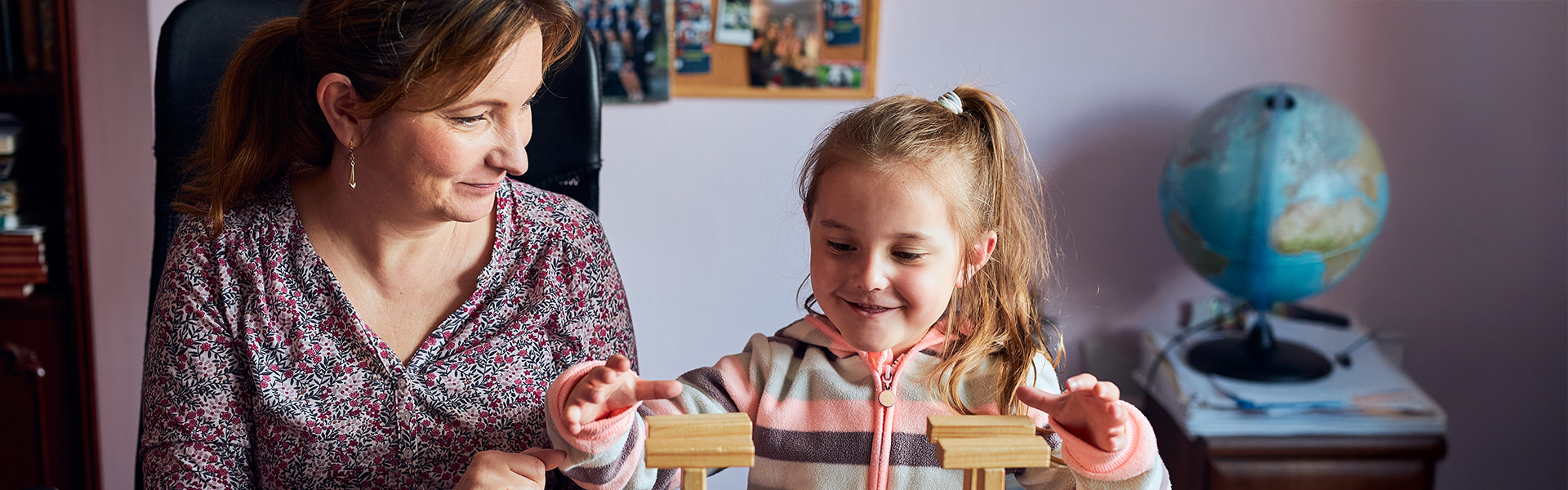 Woman sits at her dining table with her laptop open while a little girl plays with wooden blocks next to her